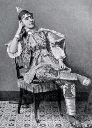woman, seated, one leg crosse at knee, wearing elaborate embroidered or woven shoes, pants, tunic and pointed hat