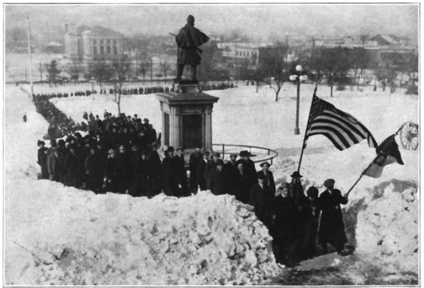woman at head of a crowded procession on a path through deep snow with a men holding a flag on either side of her